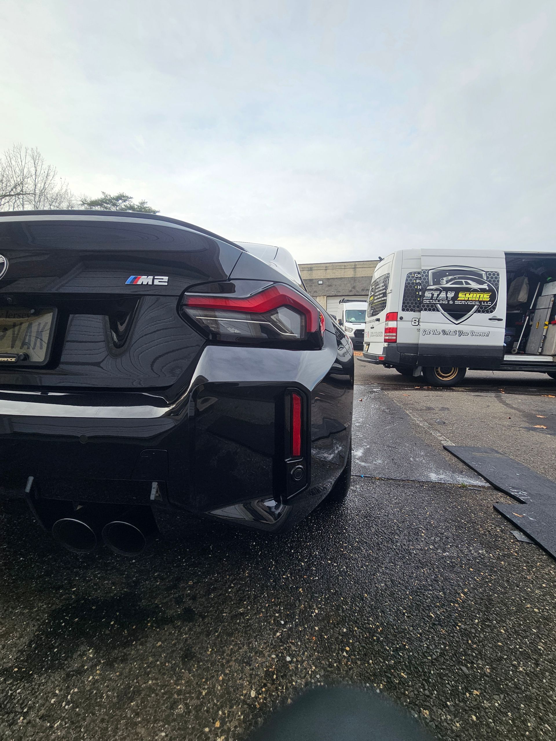 Black BMW M2 rear view, wet asphalt, parked near a van on a cloudy day.