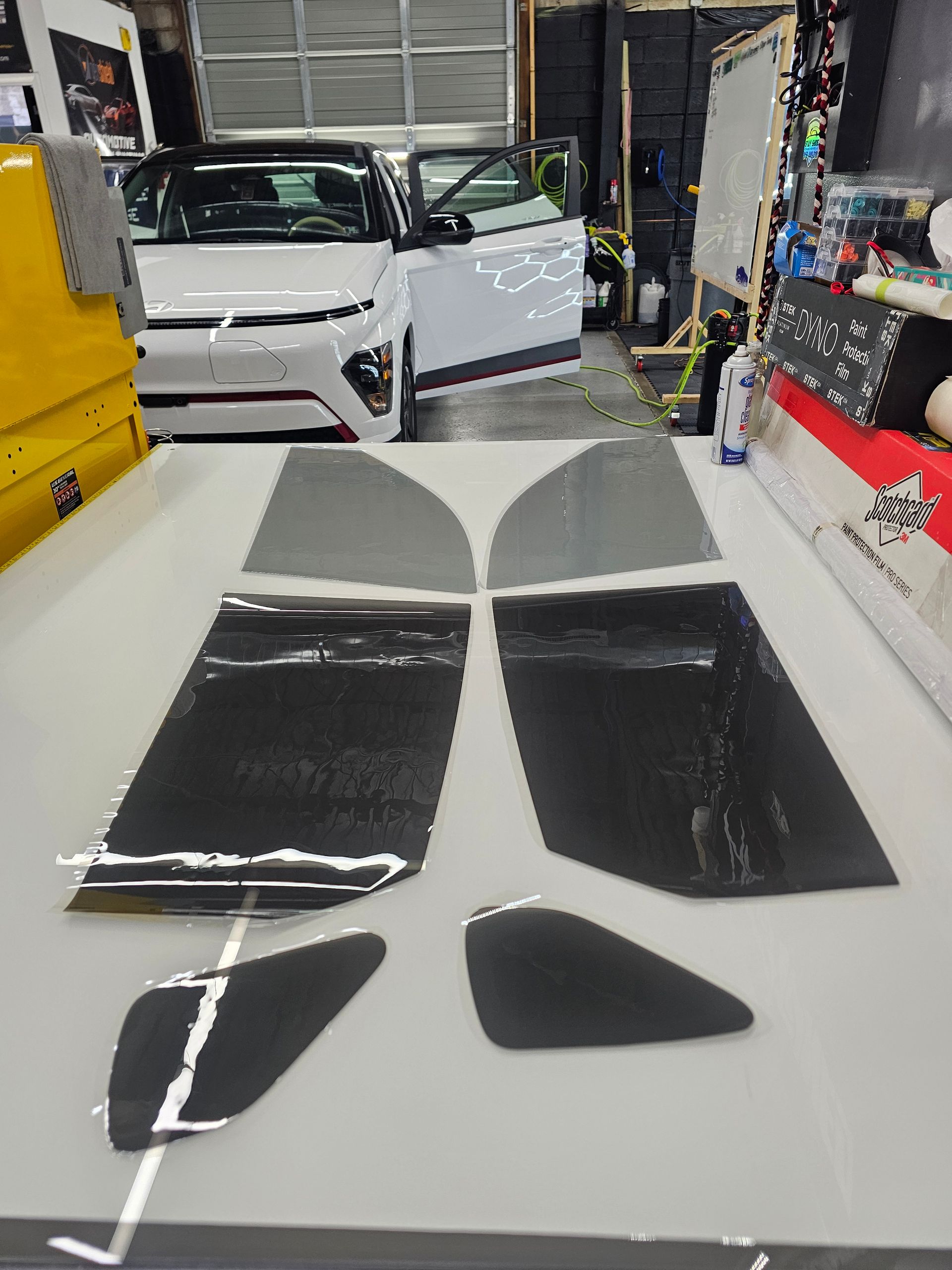 Window tinting film cutouts on a table, with a white car and open door in the background.