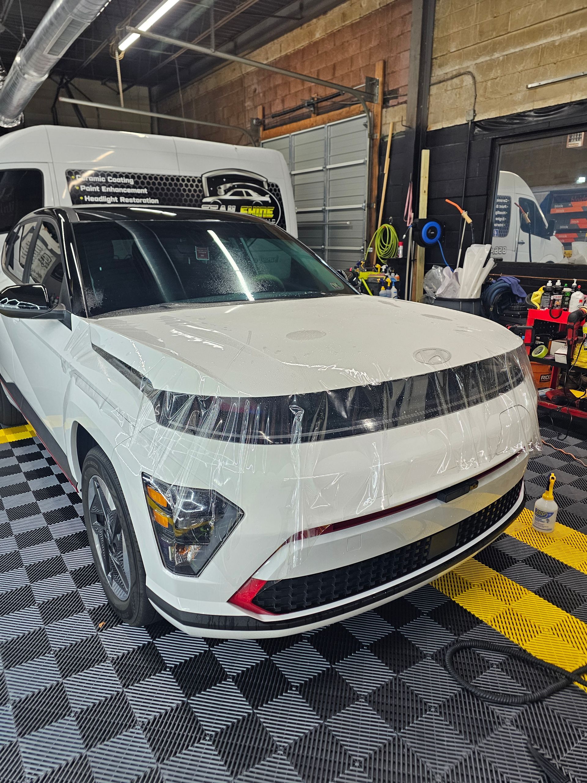 White car in a garage with protective film, black roof and trim. Black and white tiled floor.