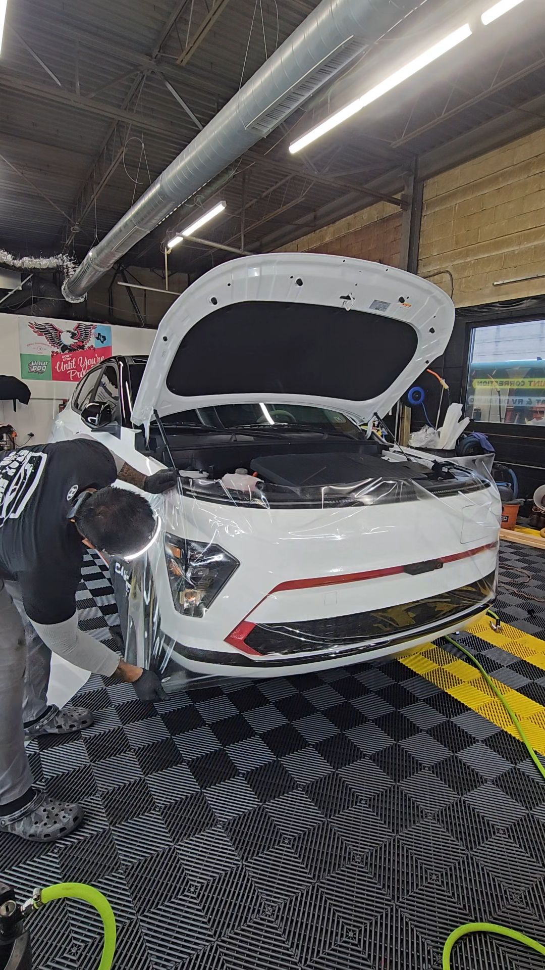A mechanic works on a white car with its hood open in a garage. Black and red trim is visible.