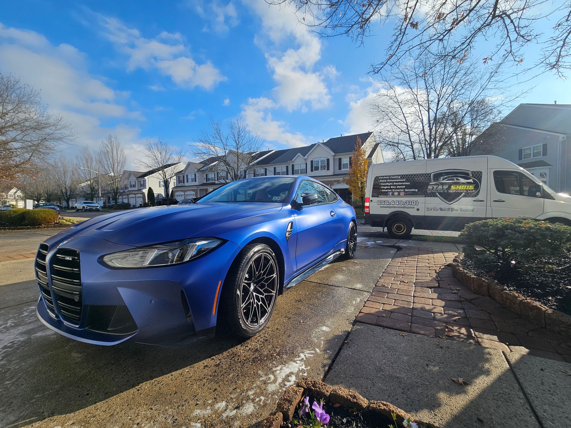 Blue BMW sports car parked on a brick driveway, next to a white van under a cloudy sky.