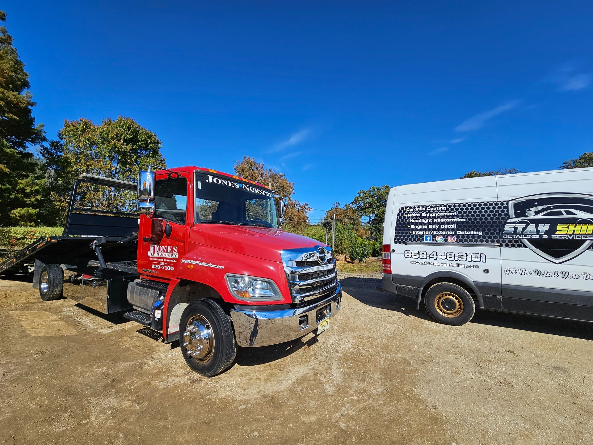 Red tow truck next to a white van with logos on a gravel lot under a blue sky.