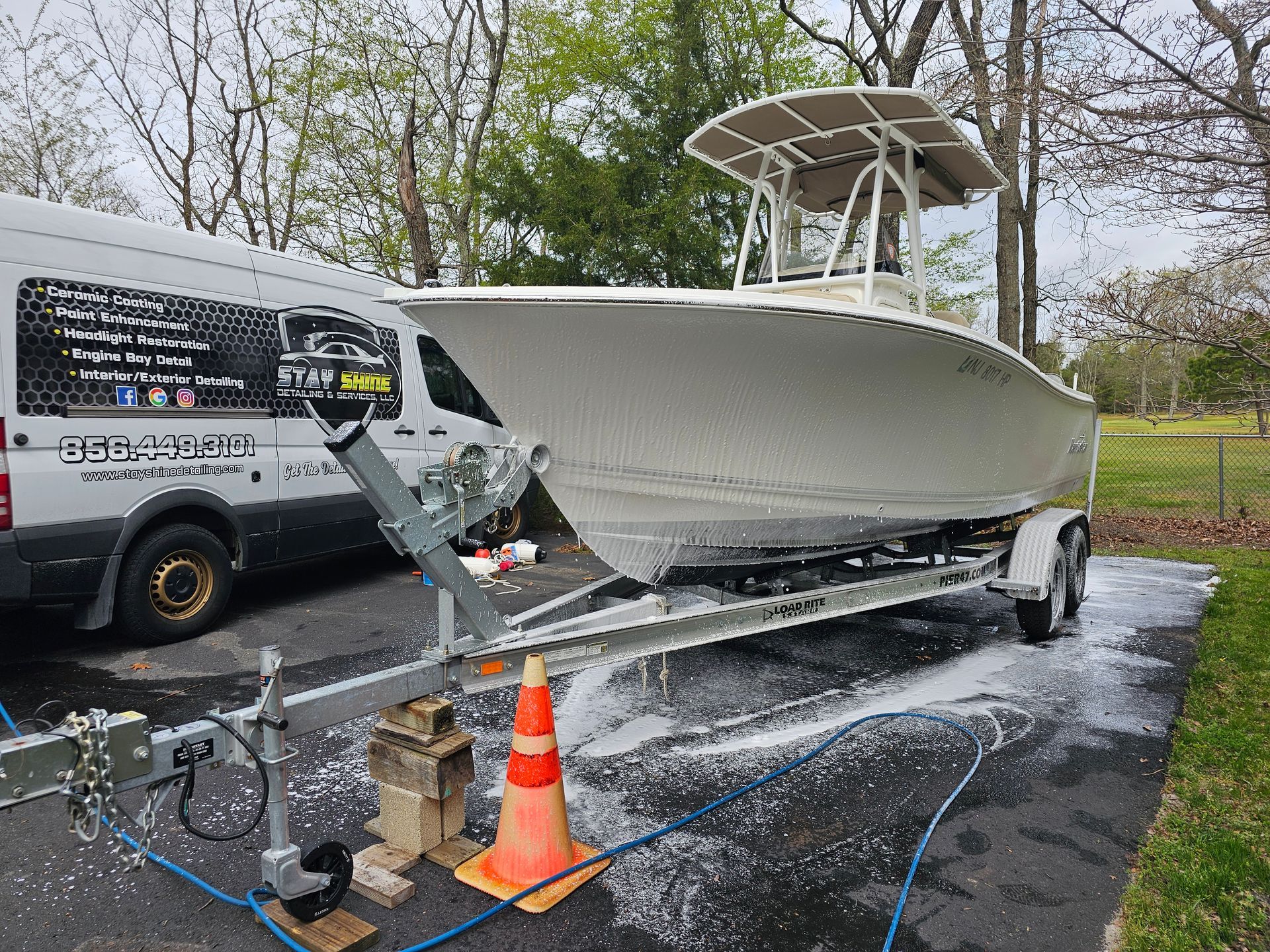 Boat being cleaned with soapy foam on a trailer next to a van. Outdoor setting, overcast.