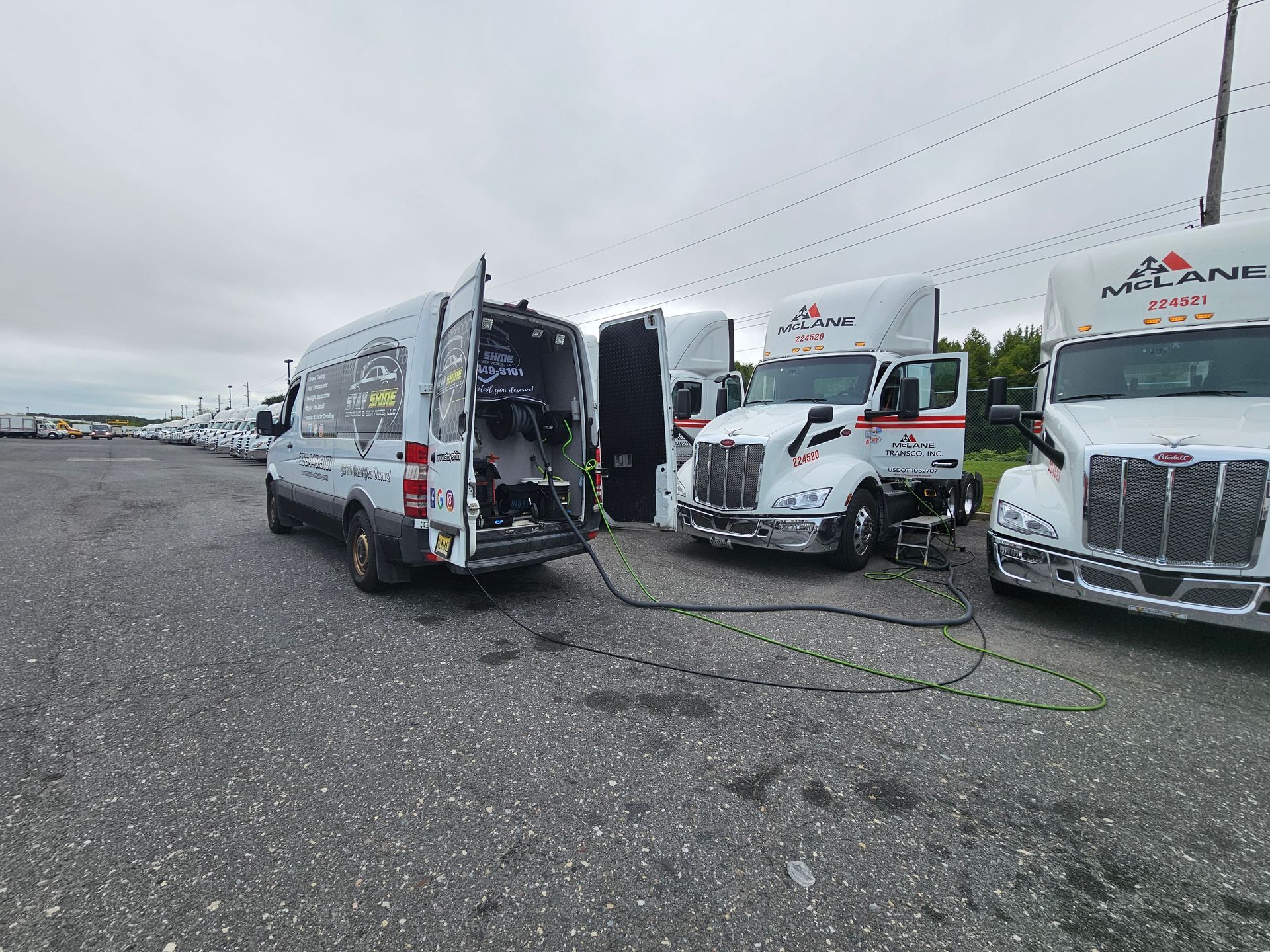 A white service van parked next to three white semi-trucks. Cables connect the van to the trucks in a paved lot.