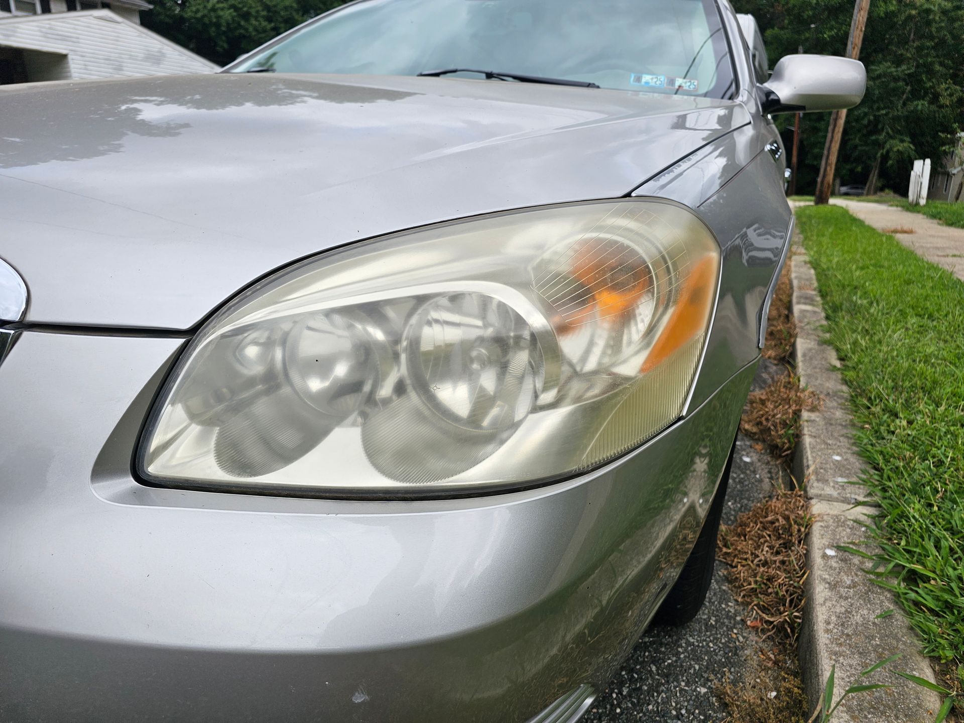 Silver car's front with cloudy headlight, parked next to a curb and grass.