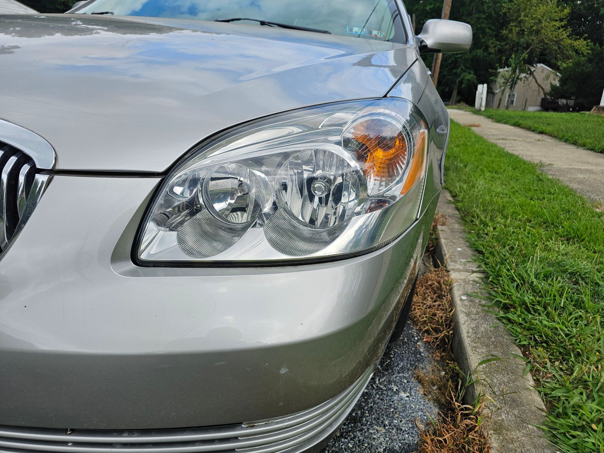 Silver car's front with headlight detail; parked on a street next to a curb and grass.