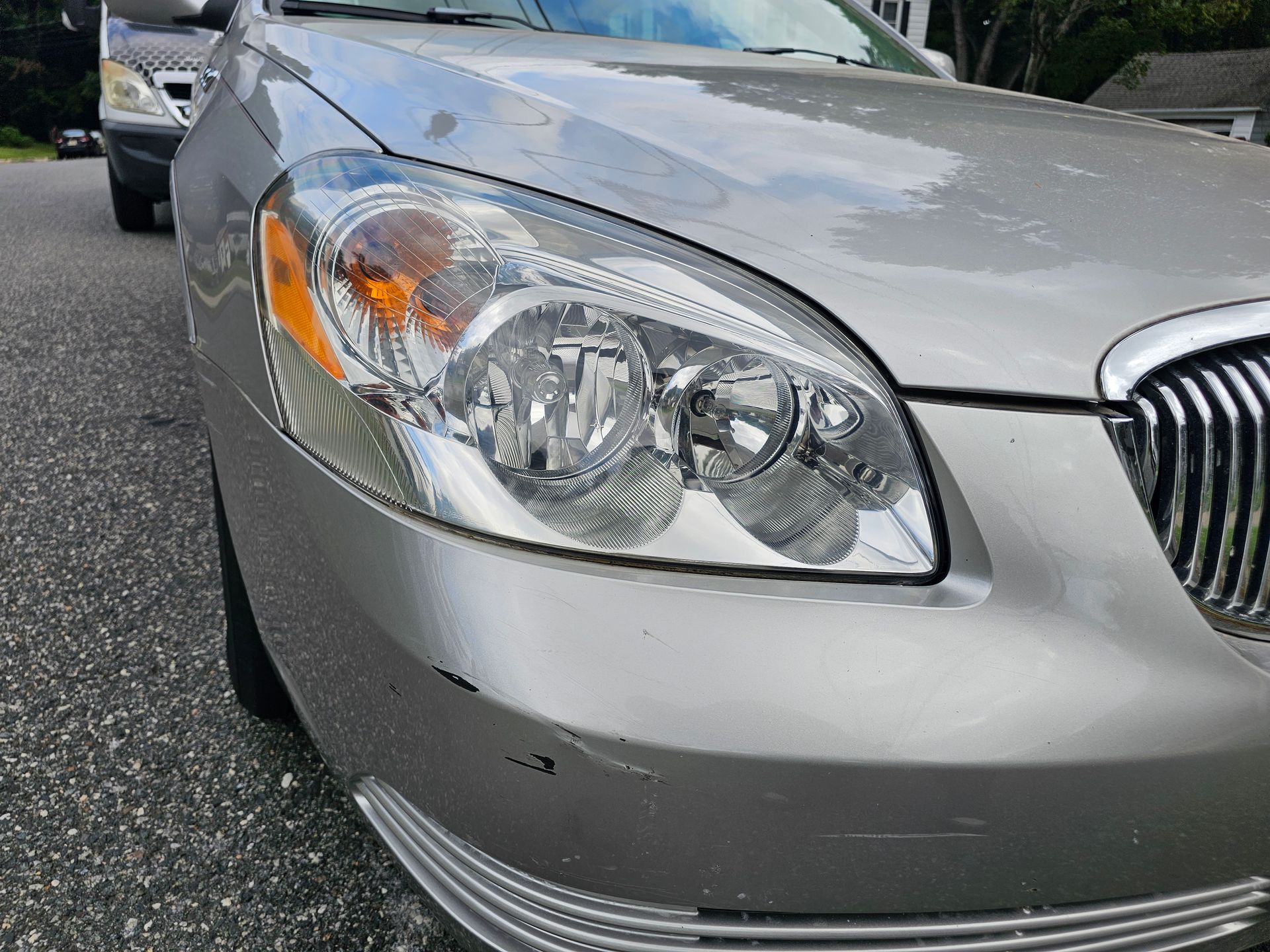 Silver Buick car's front with headlight and grill visible; parked on a road.