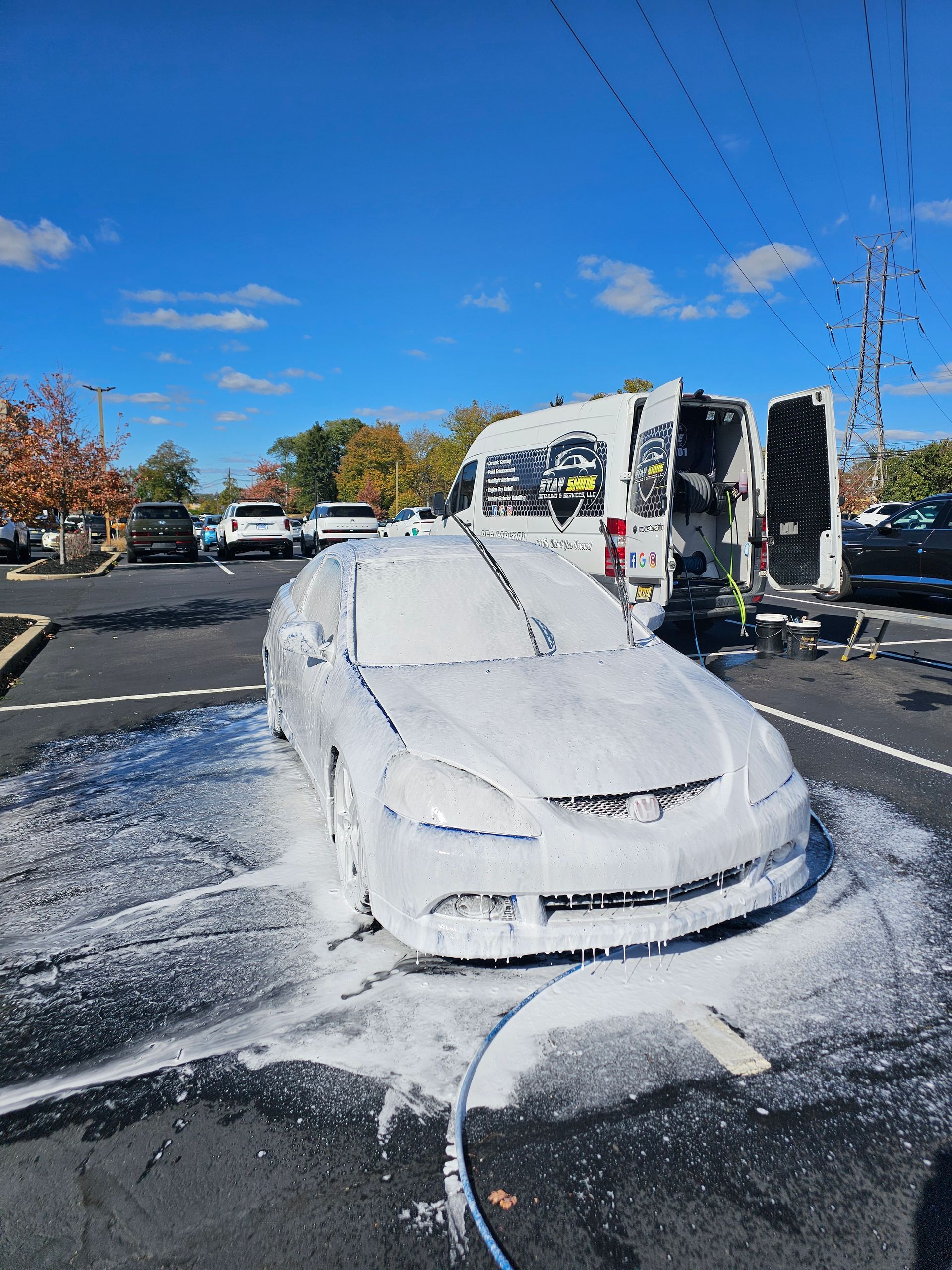 Car covered in white foam being washed outdoors, with a van and blue sky in the background.