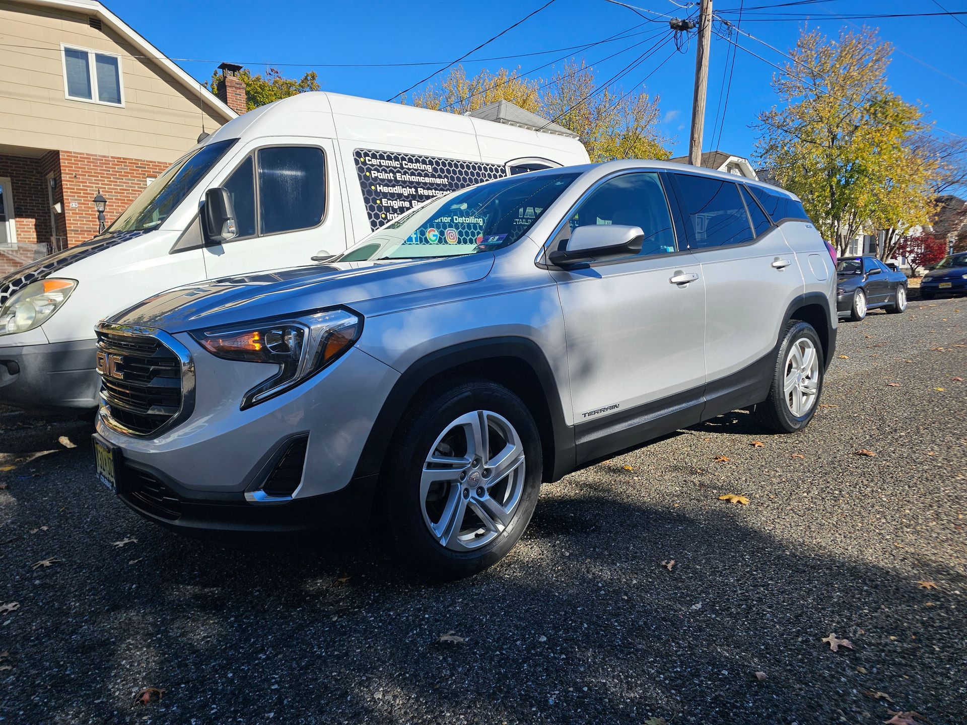 Silver GMC Terrain parked on a paved lot beside a white van and a residential building on a sunny day.