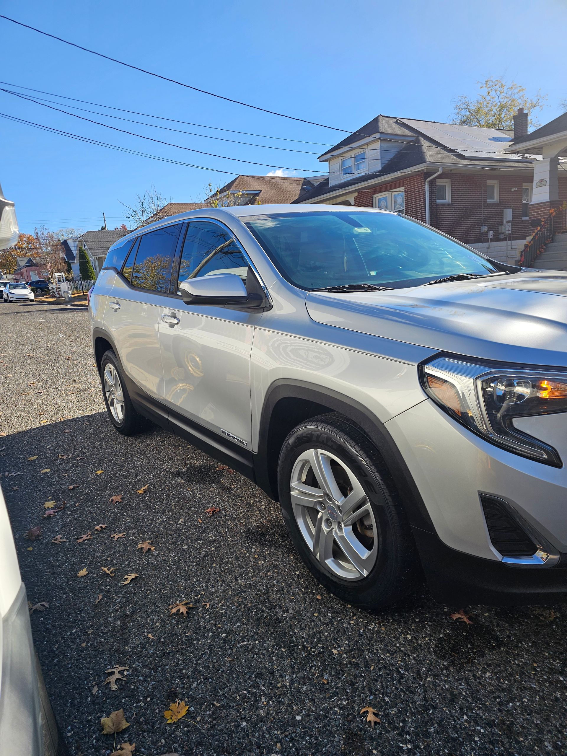 Silver GMC Terrain parked on a gravel surface next to a road, houses in background under a blue sky.