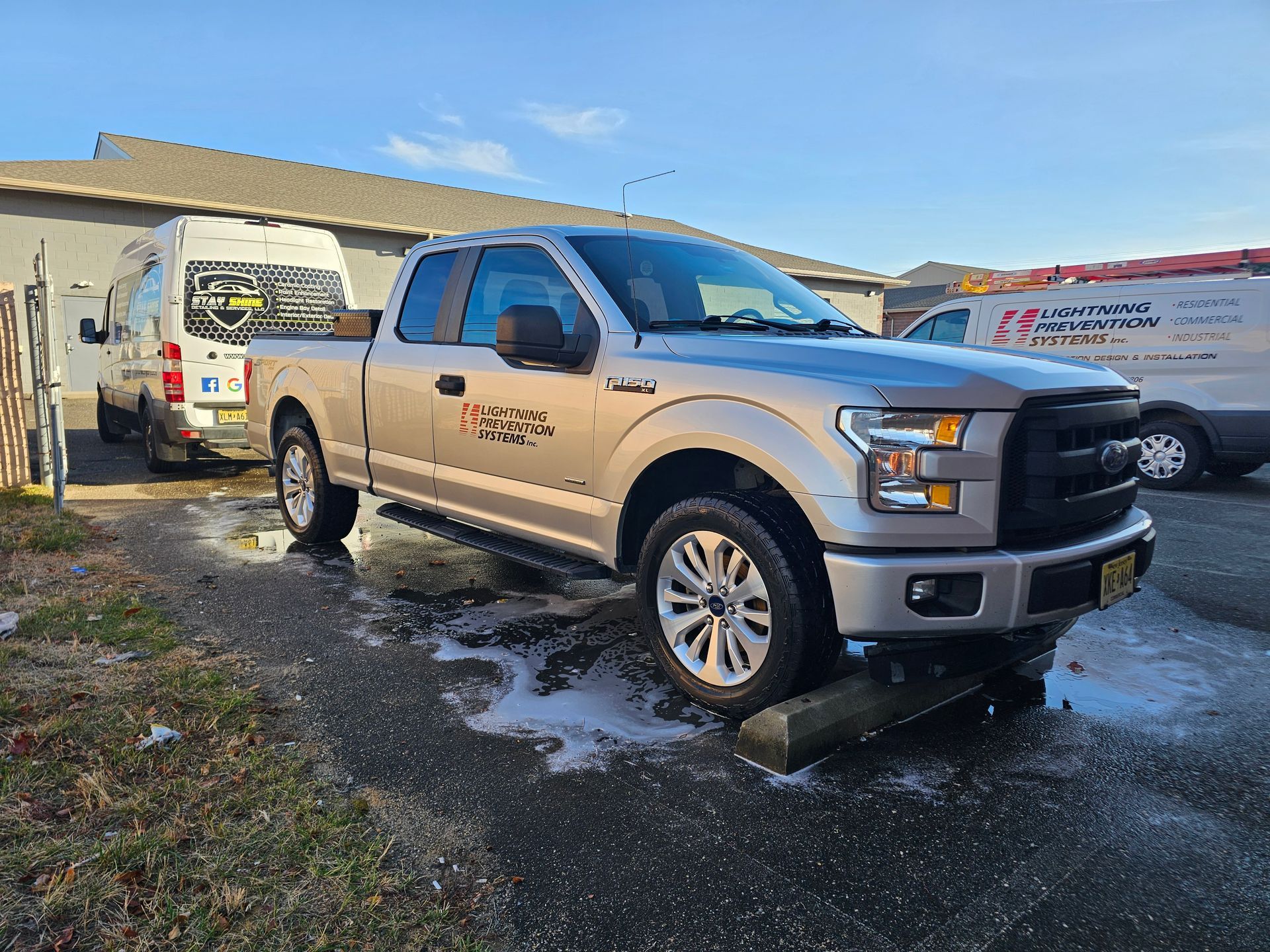 Silver Ford pickup truck parked on concrete blocks next to a white van with business logos on a sunny day.
