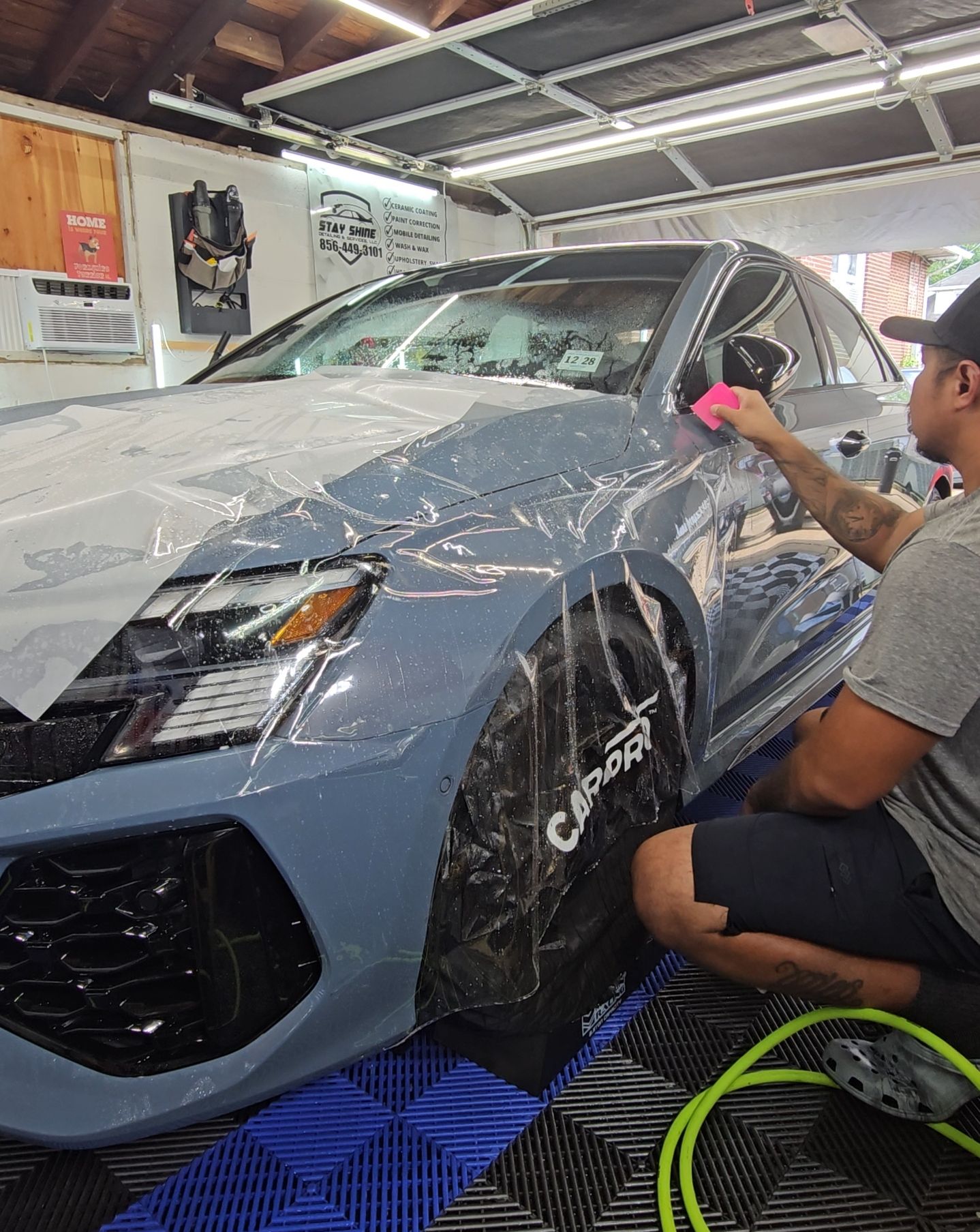 Man applying protective film to a gray car in a garage, using a squeegee.