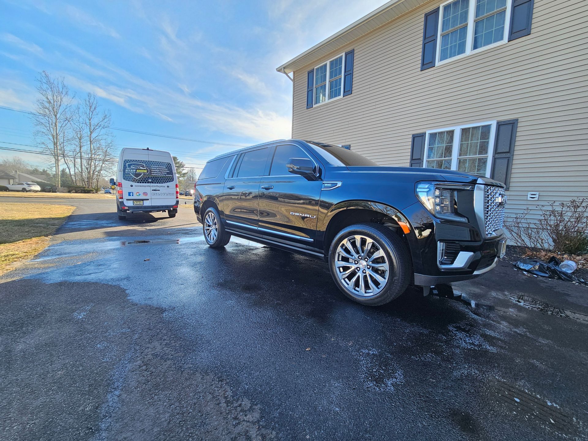 Black GMC Yukon SUV parked on wet pavement in front of a tan two-story building on a sunny day. A van is in the background.