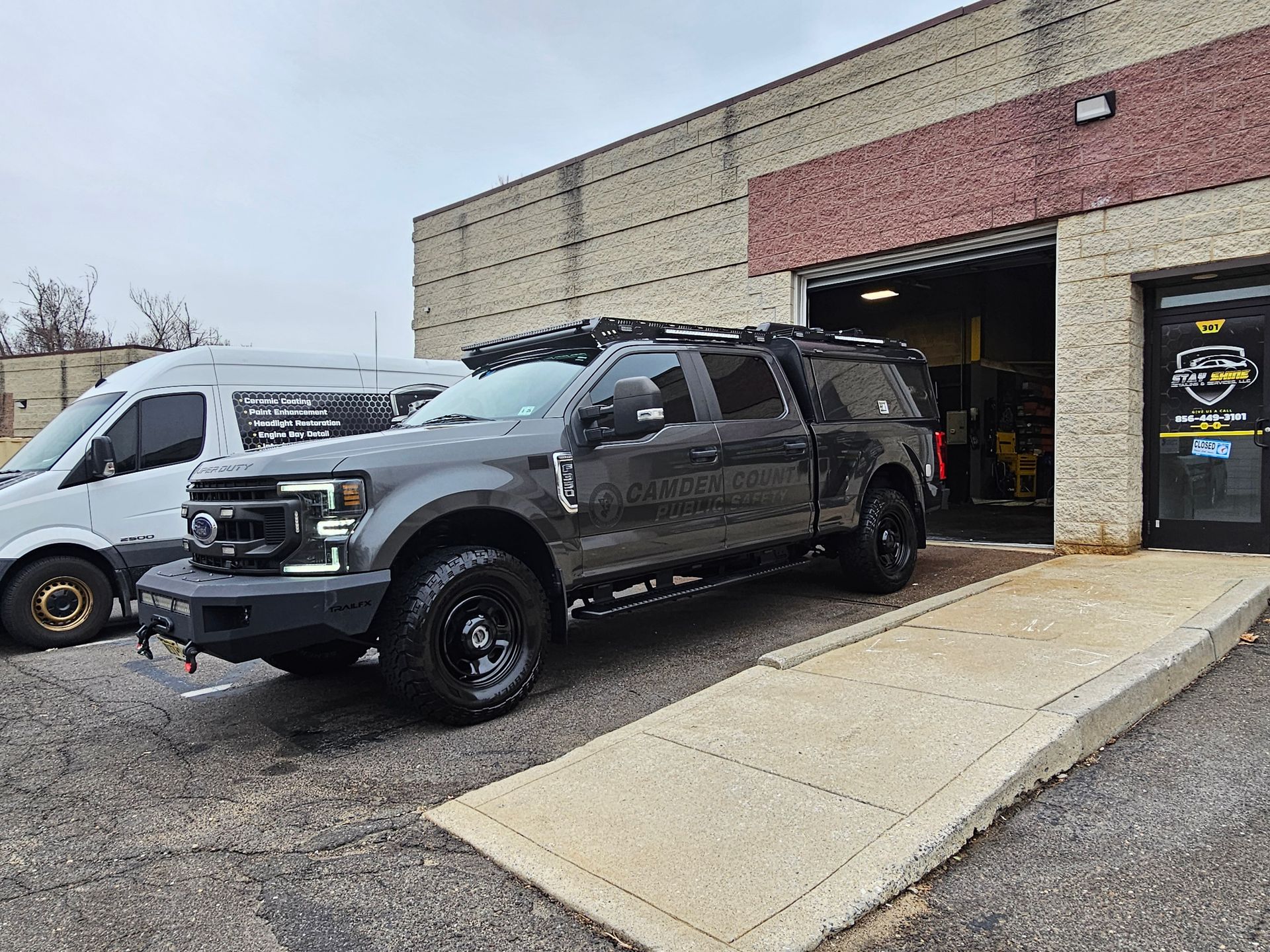 Dark gray pickup truck with black wheels and a roof rack parked outside a brick building with open garage door.