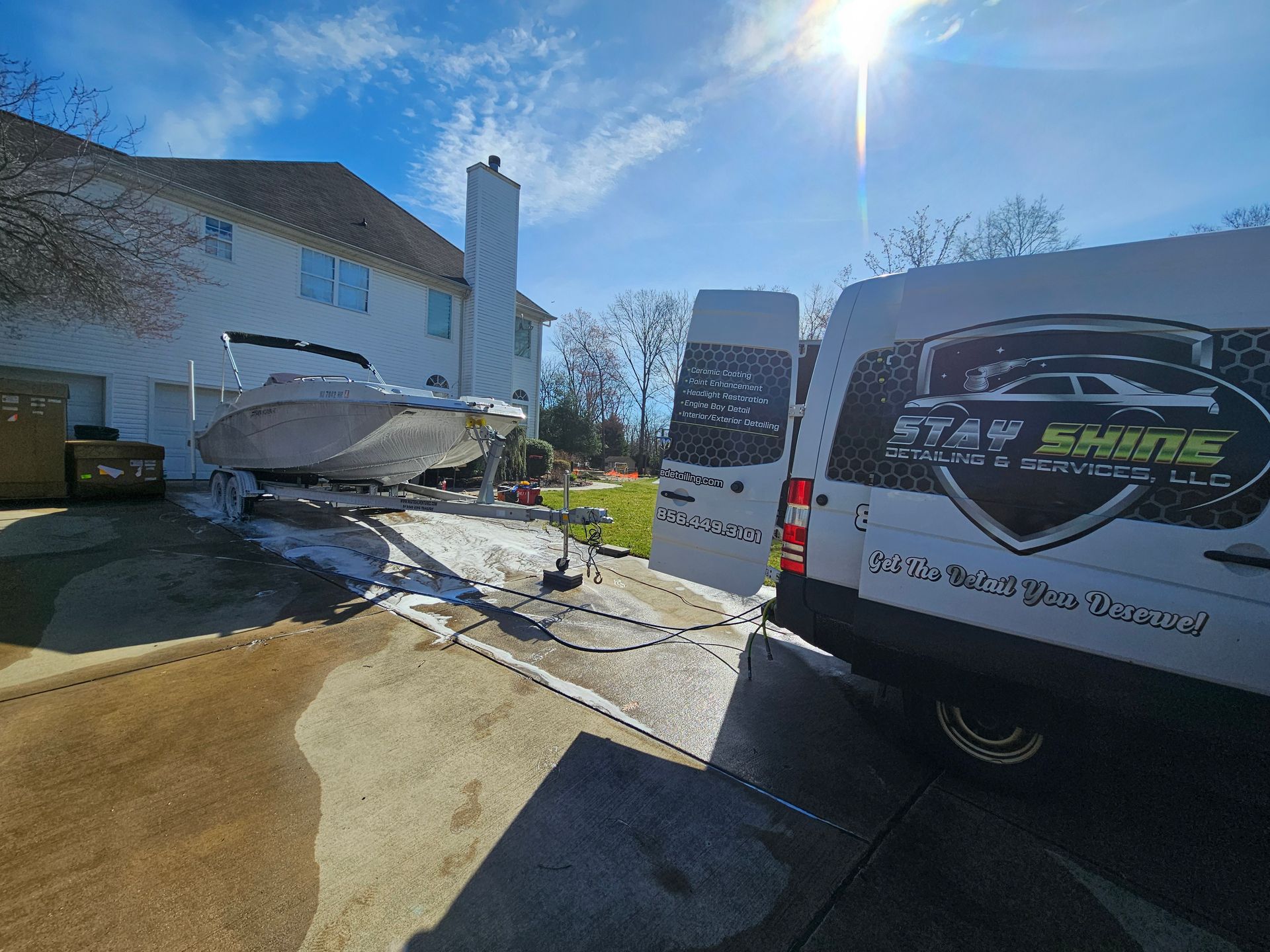A boat on a trailer being washed by a detailing van in a driveway on a sunny day.