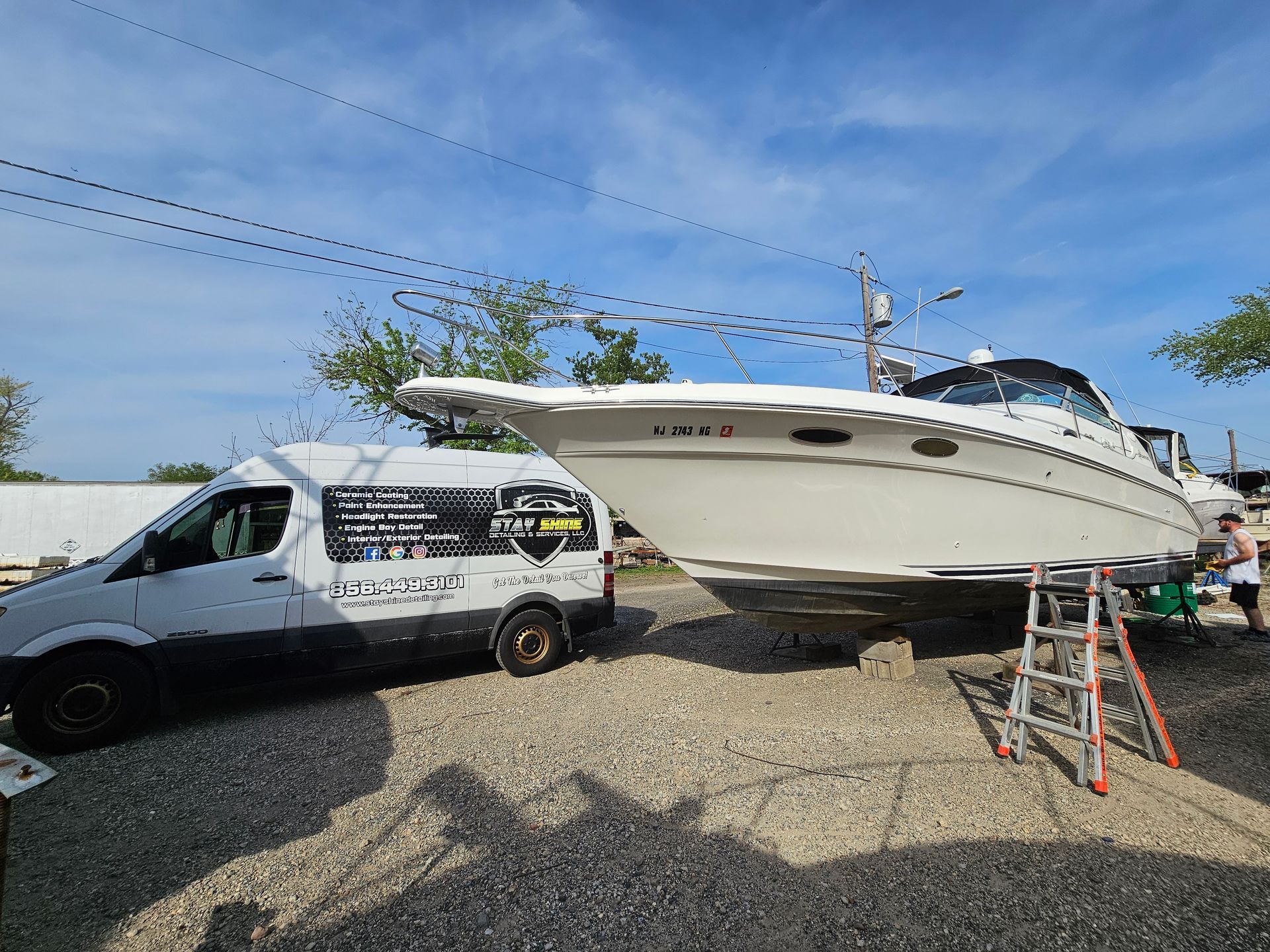A white boat on blocks next to a white van in a gravel lot under a blue sky.