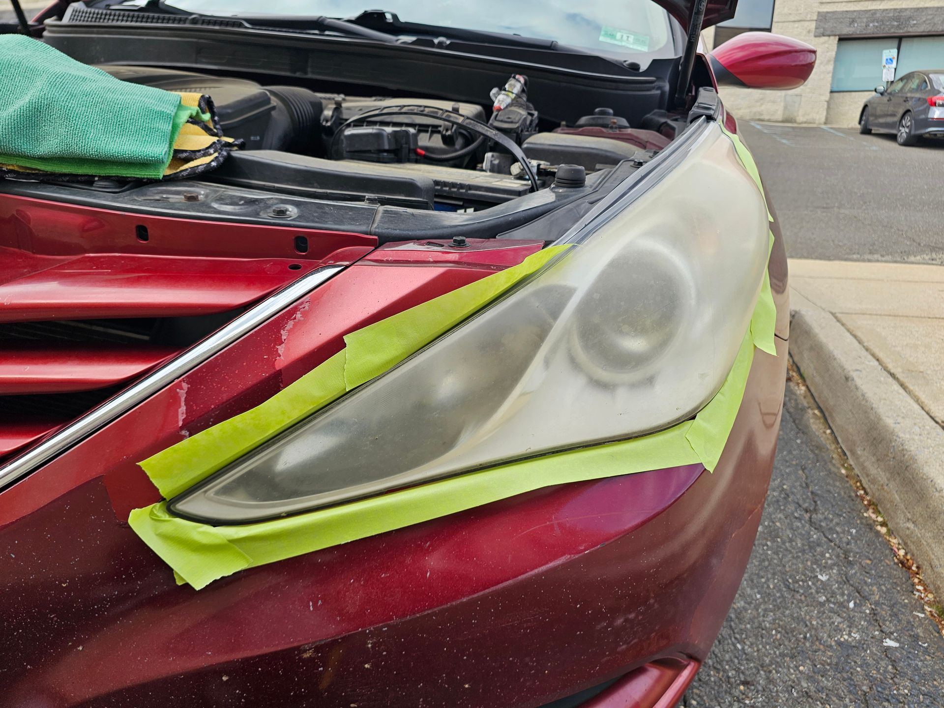 Car headlight being restored, masked with green tape, on a red car with the hood open.