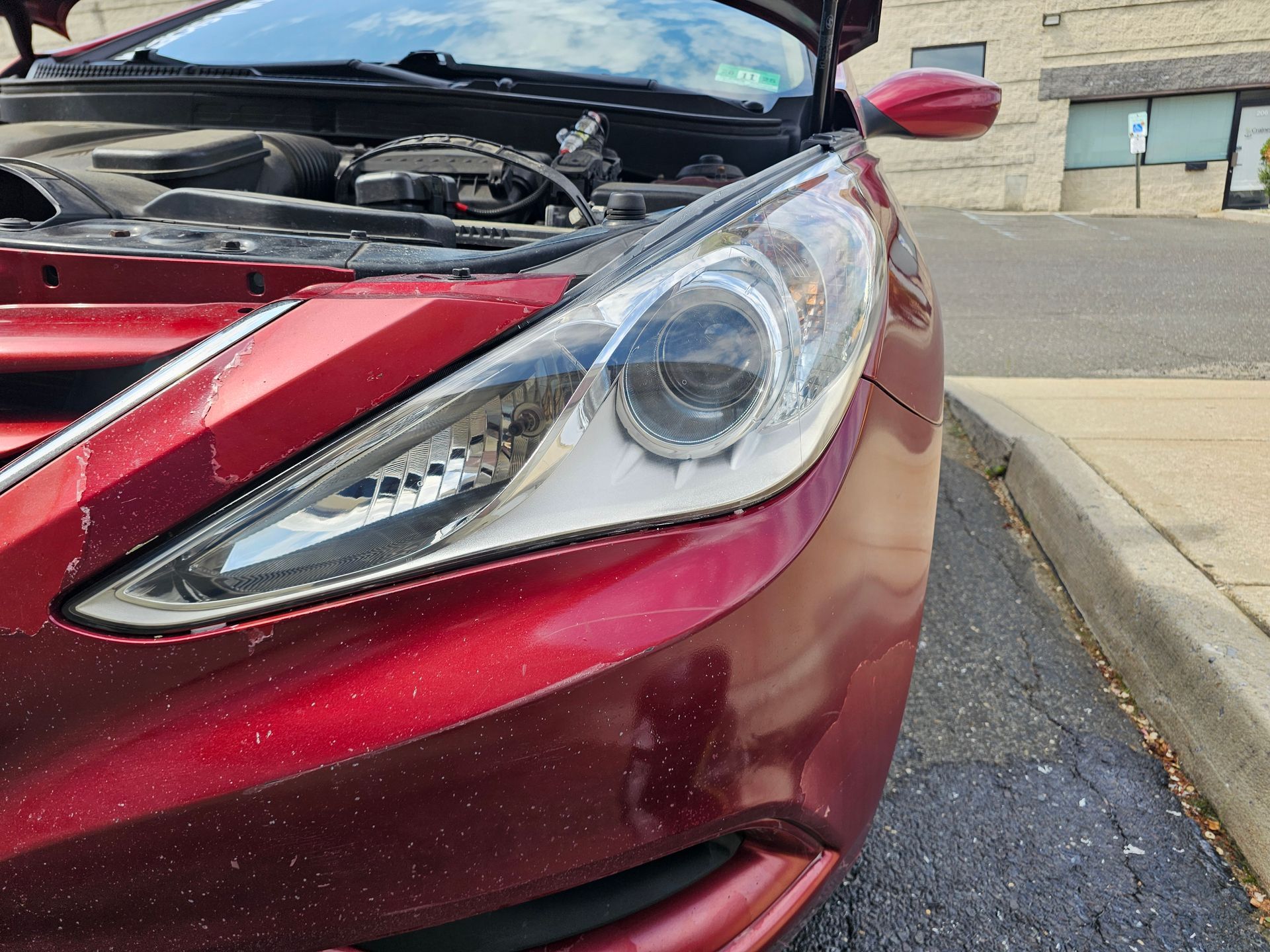Red car with hood open, showing headlight and engine. Outside, next to a curb.