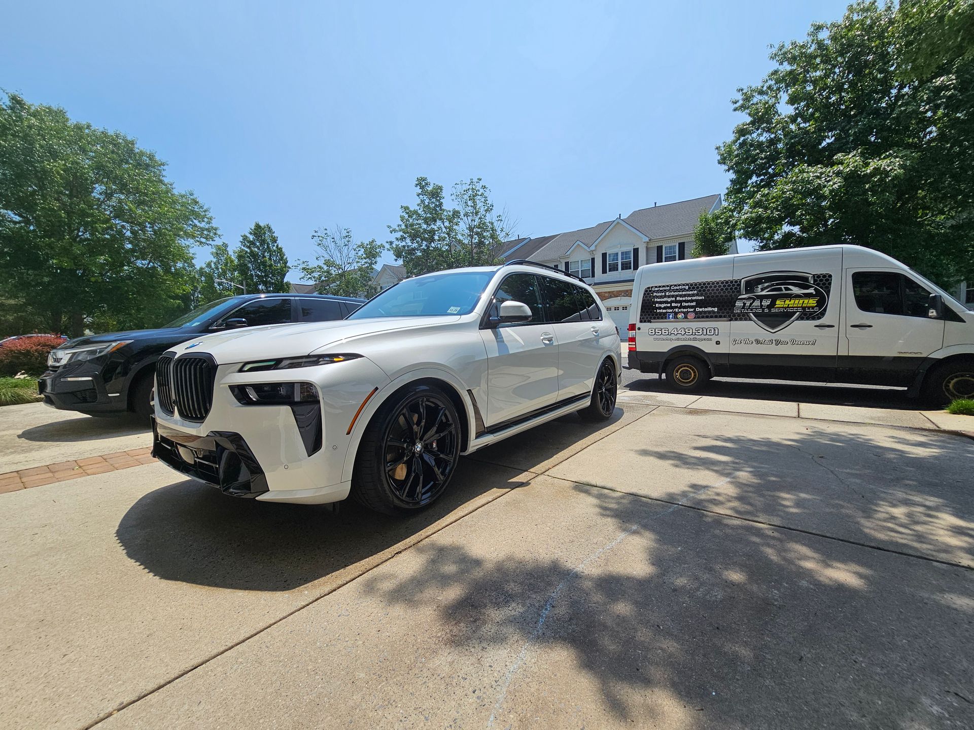 A white BMW SUV and black car parked in a driveway next to a white van on a sunny day.