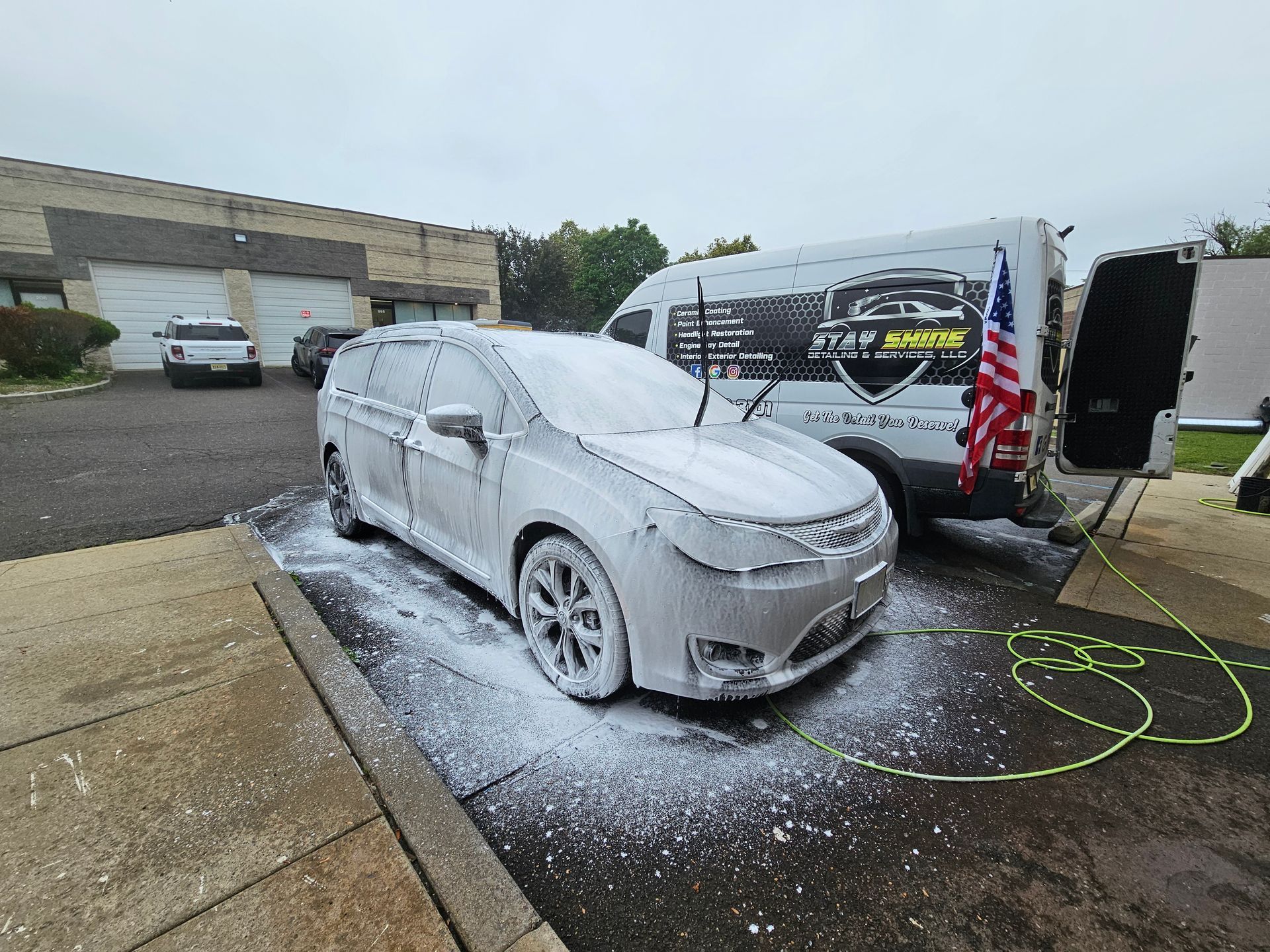 Silver minivan covered in foam being washed outside; white van with logo and American flag visible.