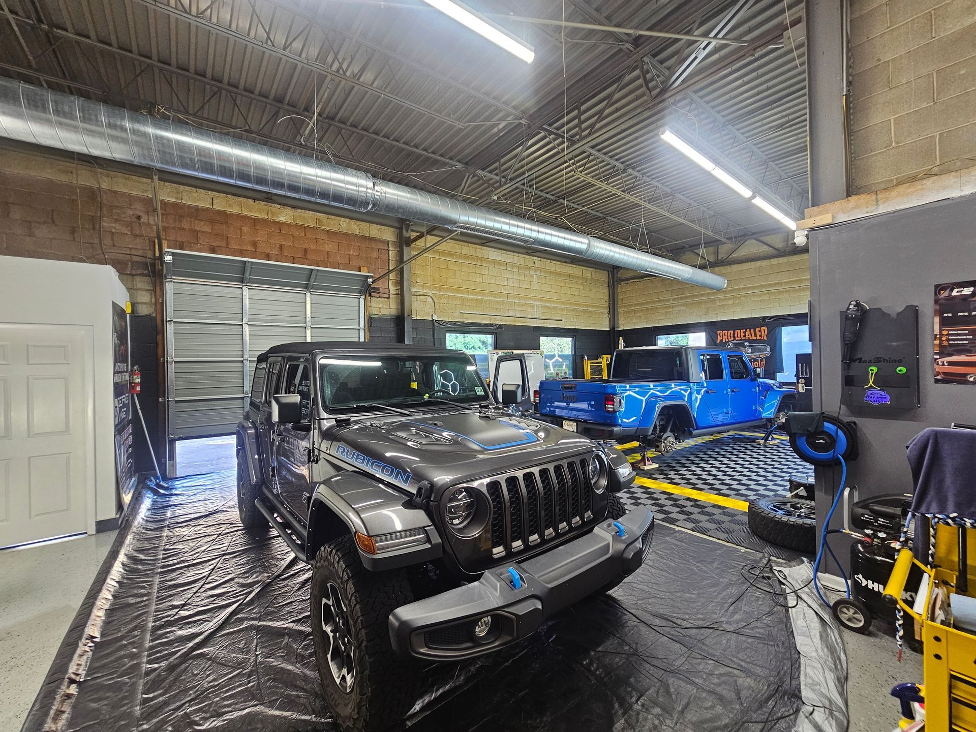 Dark gray Jeep inside a garage, with blue truck in background. Black mats on the floor, overhead lights.