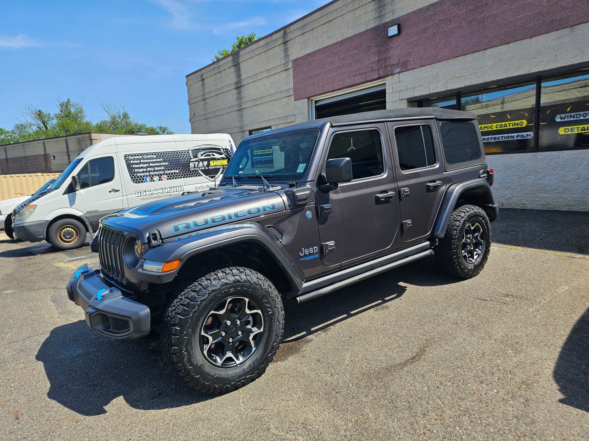 Dark gray Jeep Wrangler parked in front of a building with a white van nearby on a sunny day.