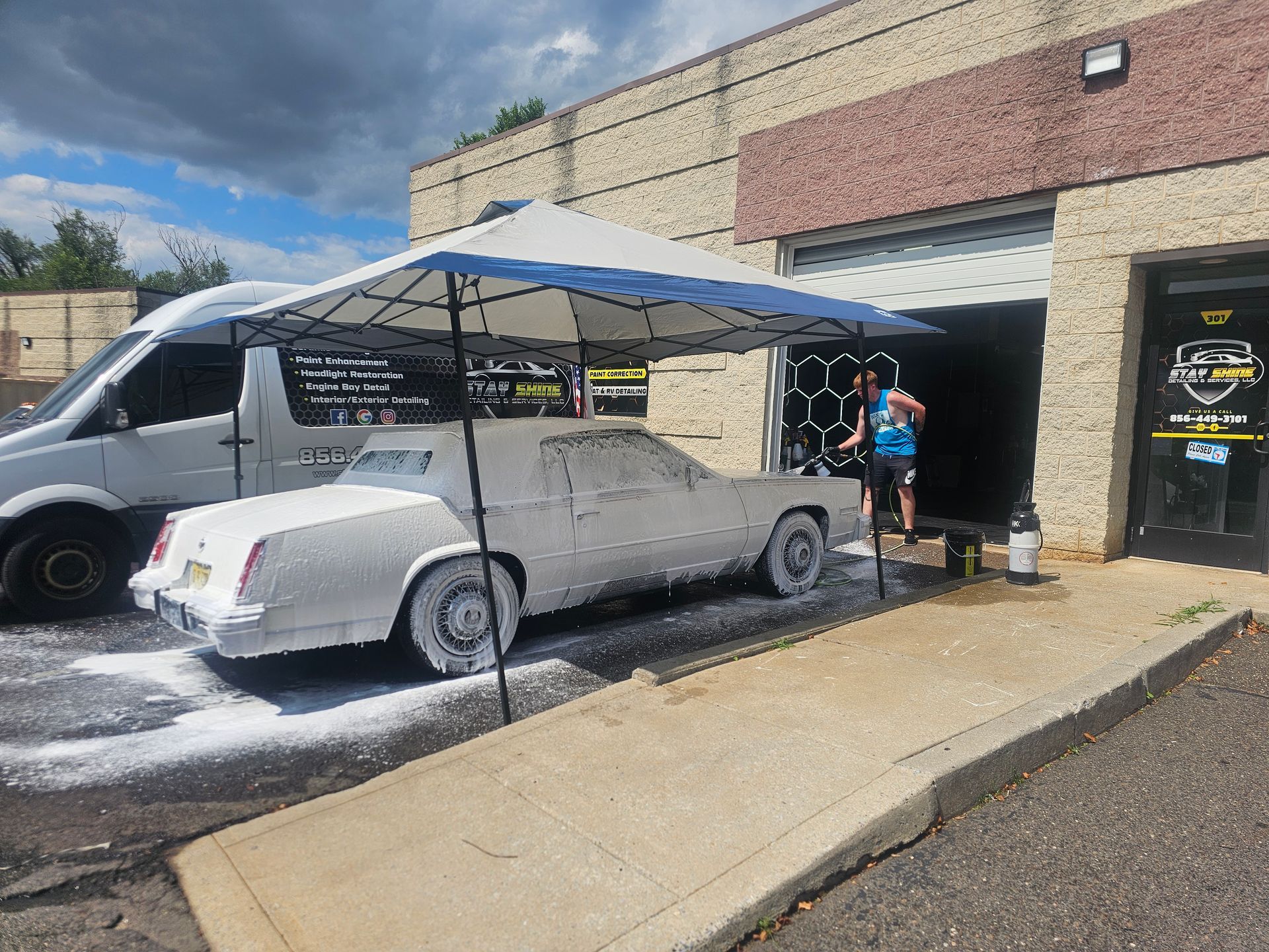 A white car covered in foam being washed outside a garage under a canopy.