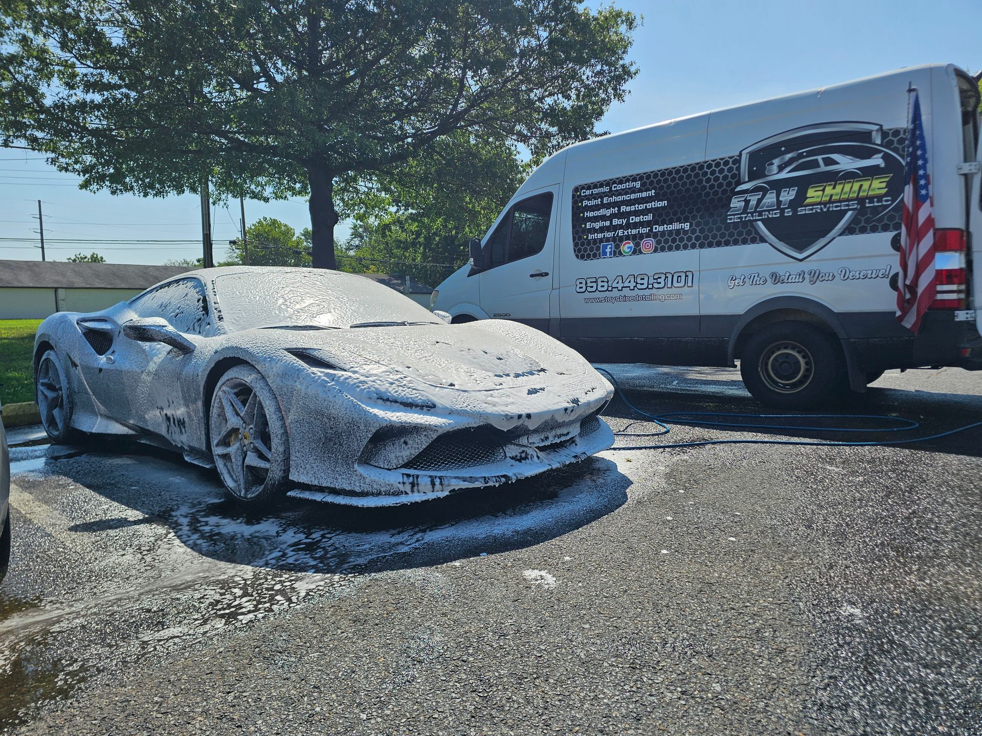 A gray sports car covered in white foam sits in a parking lot. A white van with graphics is behind it.