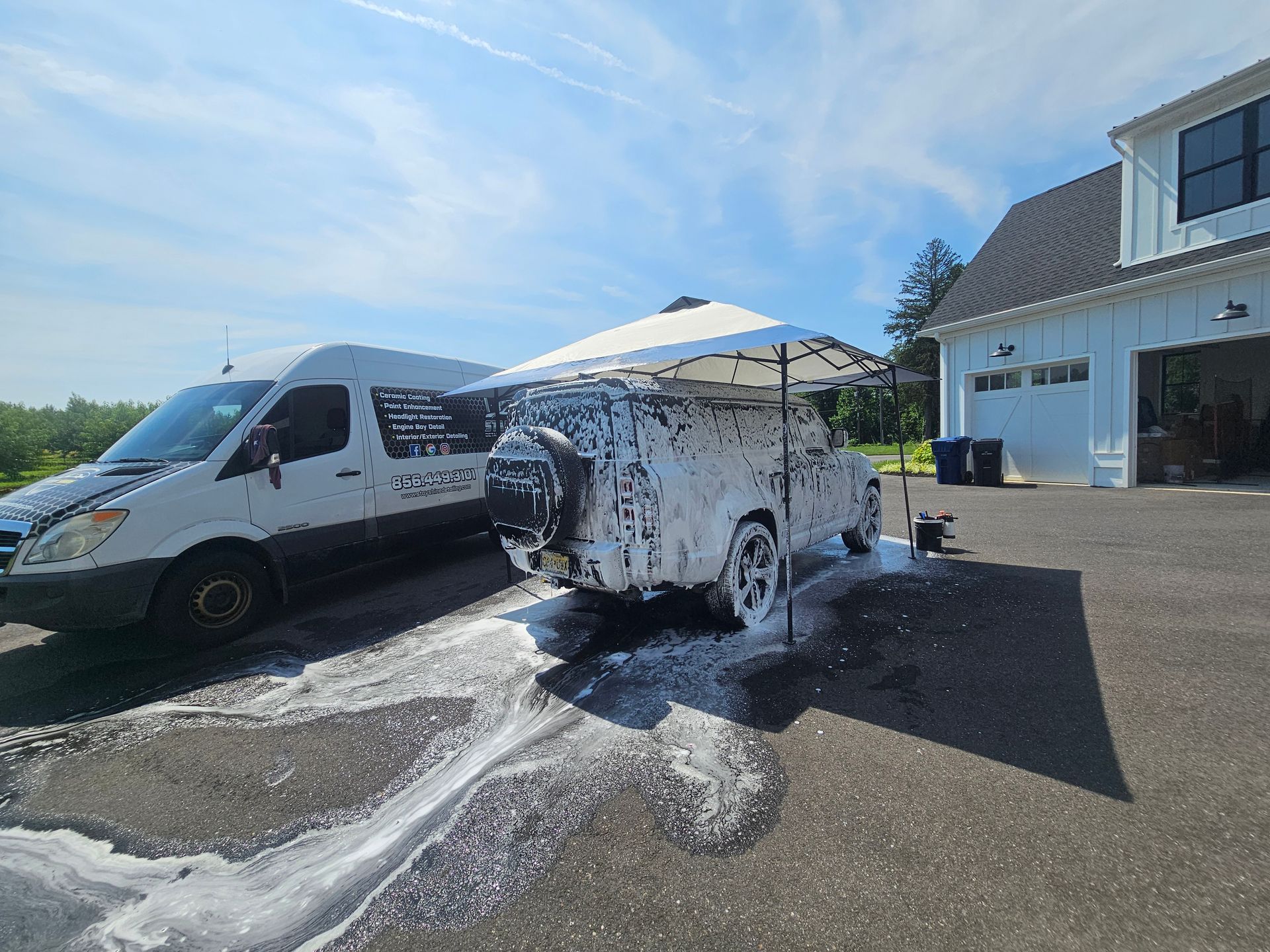 White car covered in foam being washed outside on a sunny day next to a van and a white building.