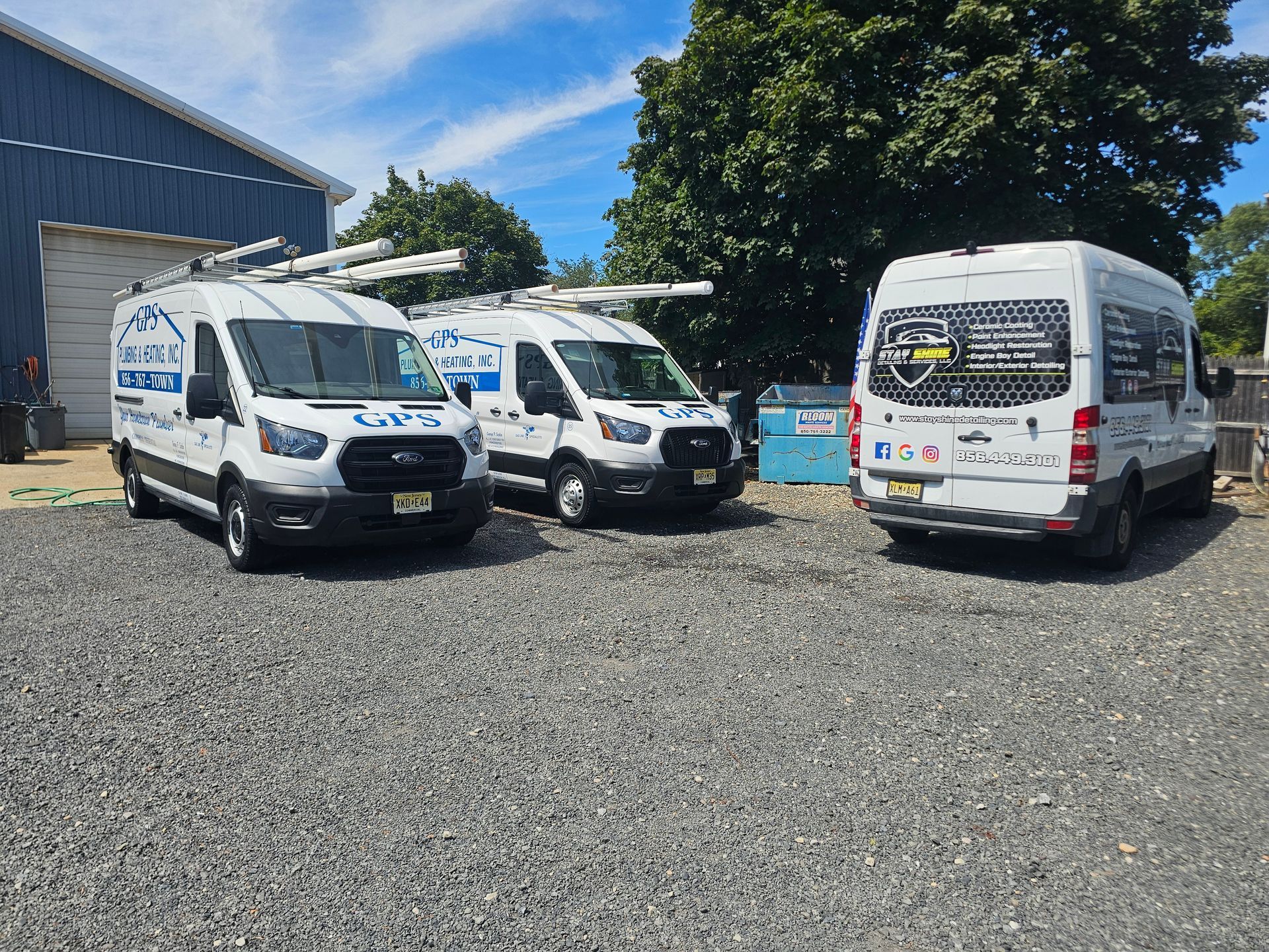 Three white service vans parked on gravel, with company logos, in front of a blue building and trees.