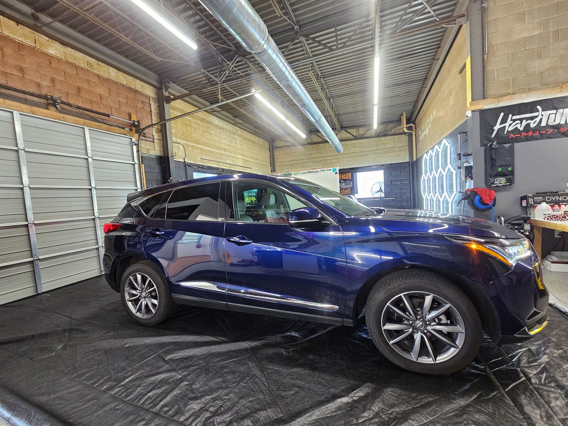 Blue Acura RDX SUV parked inside a garage. The vehicle sits on a black mat. Garage has a metal ceiling.