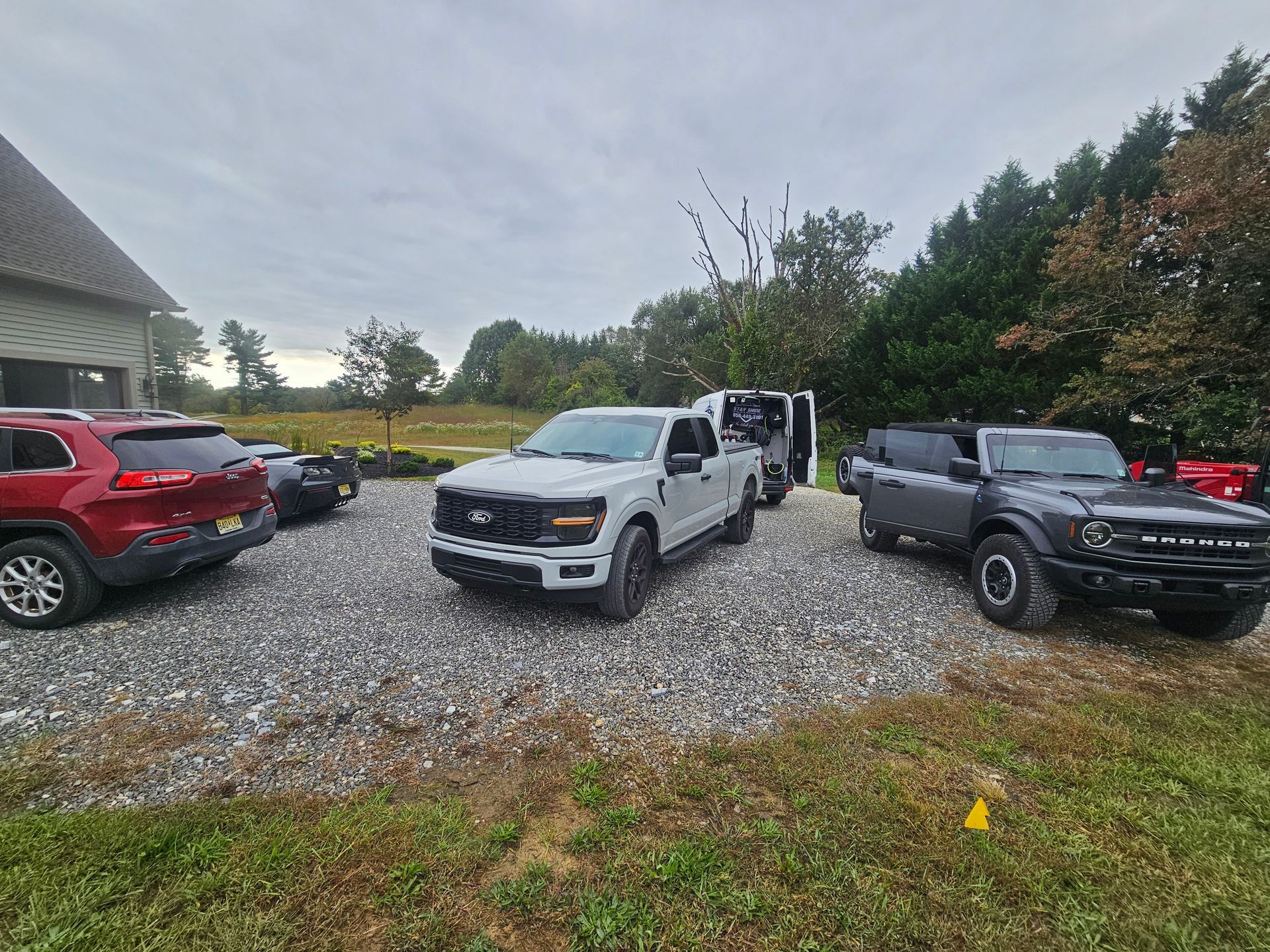Several vehicles parked on gravel: SUV, pickup truck, and a Bronco, in front of a building and trees on a cloudy day.