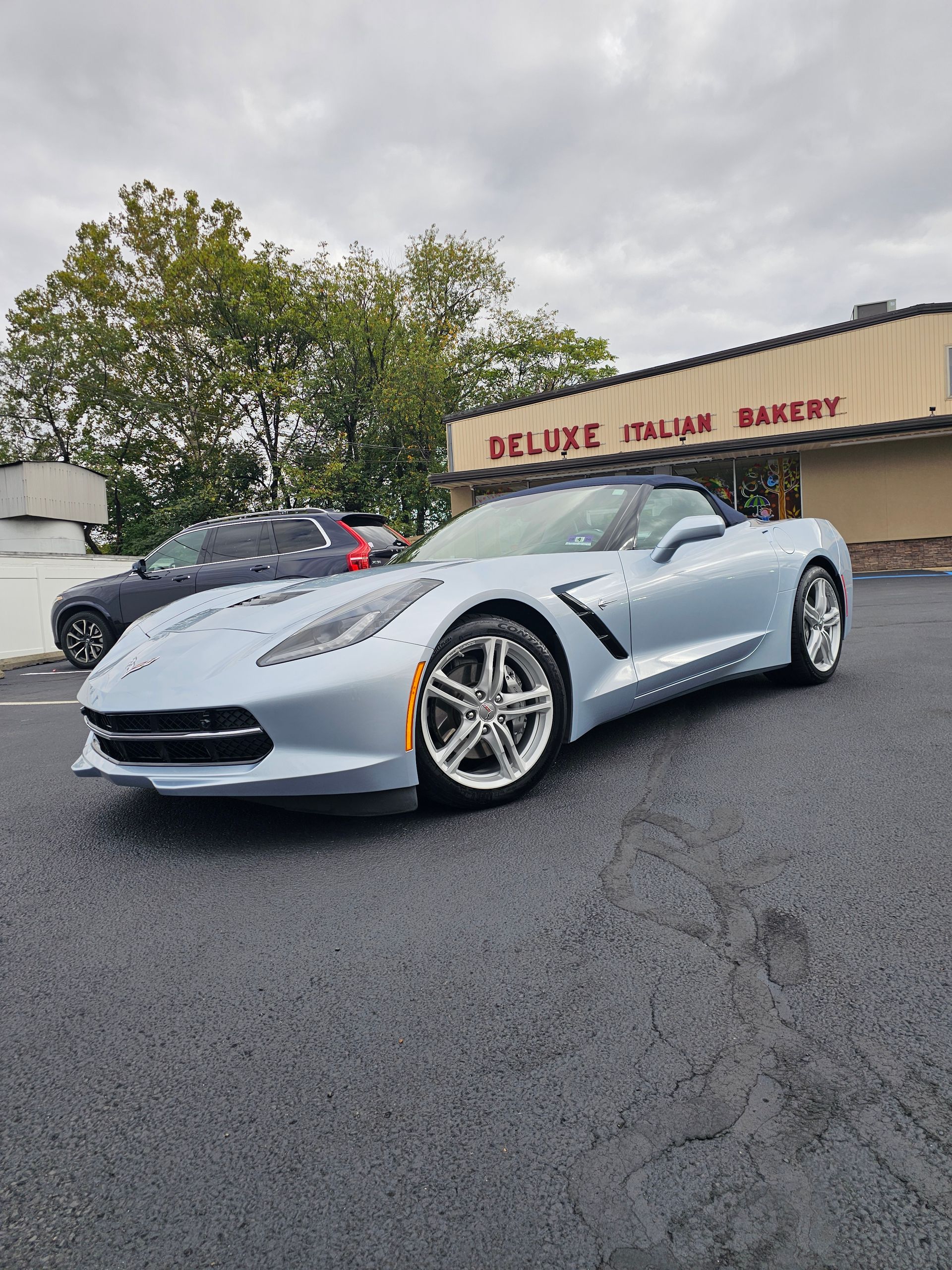 Silver convertible Corvette parked outside a bakery under a cloudy sky.