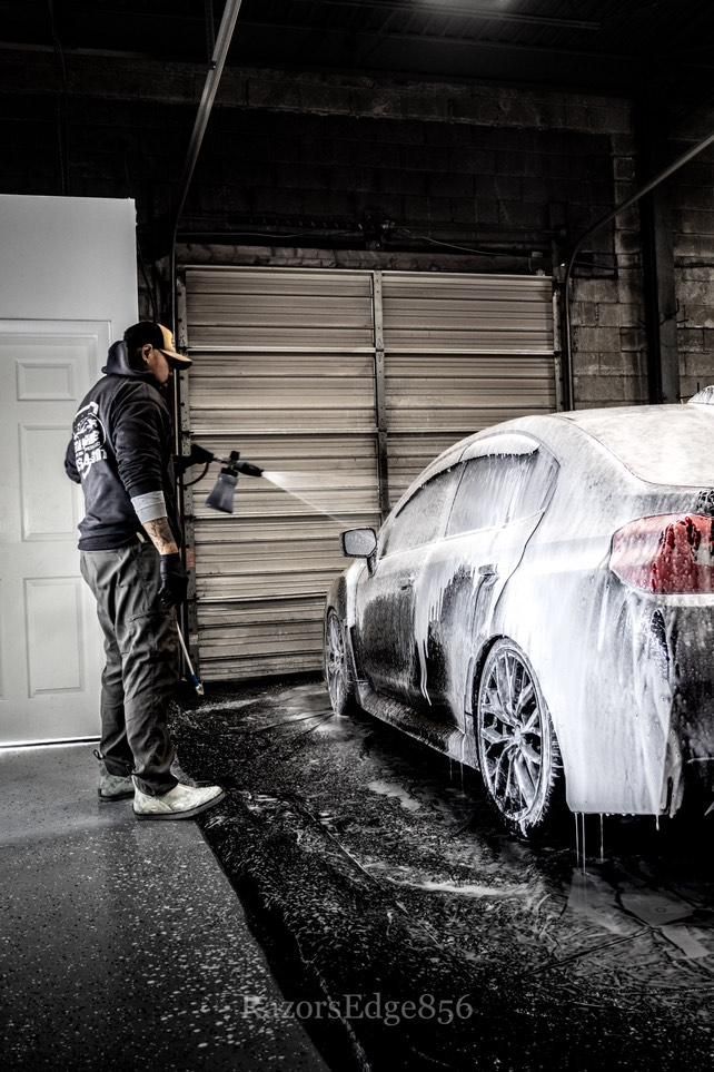 Person washes a white car in a garage with foam. They are holding a pressure washer.