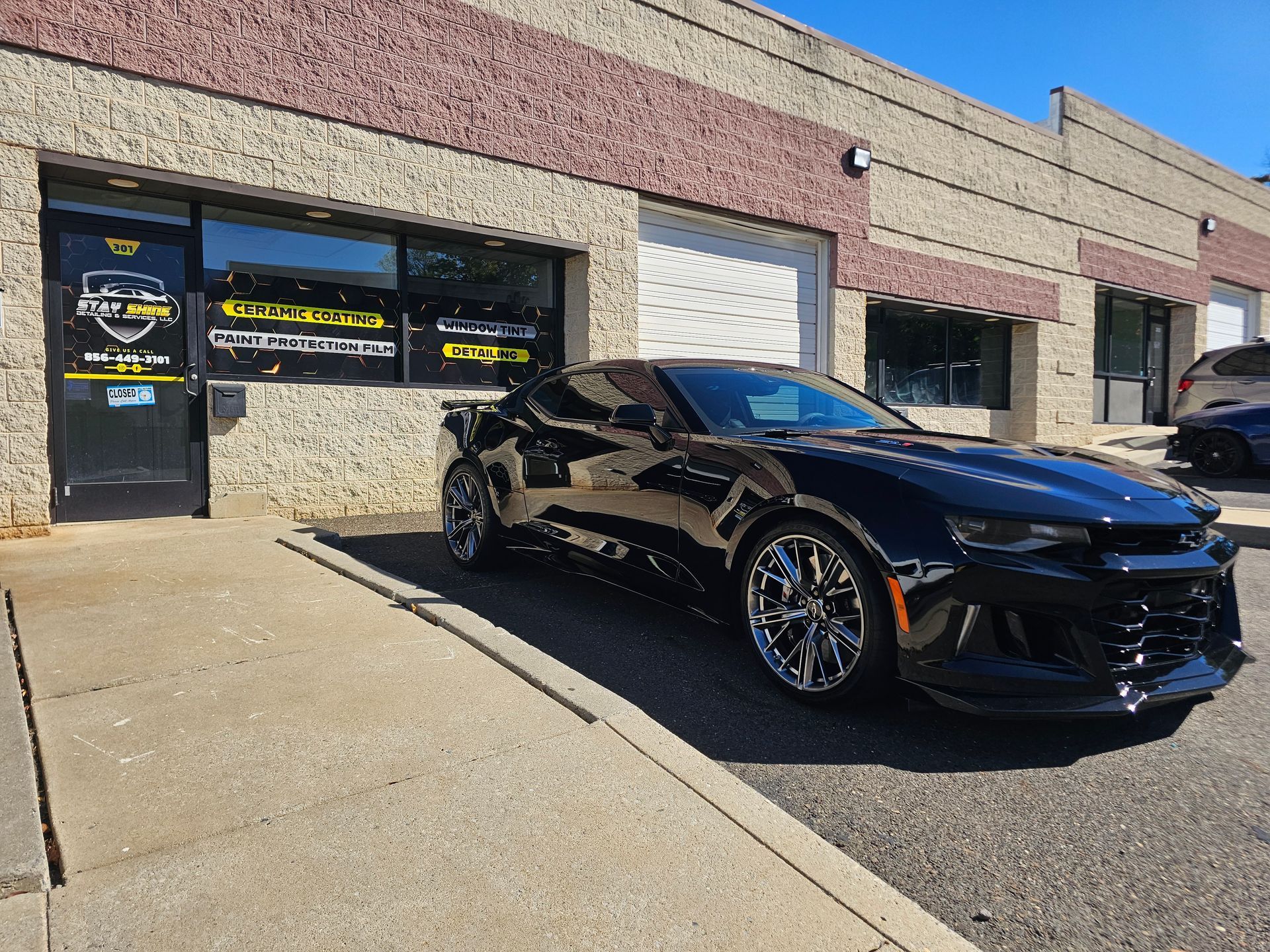 Black Camaro parked in front of an auto shop. Building is tan and gray. Bright sunny day.