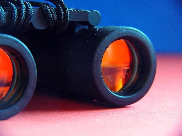 Black binoculars with orange lenses on a pink surface against a blue background