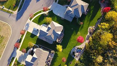 Aerial view of several houses with gray roofs, green lawns, and curving roads lined with trees.