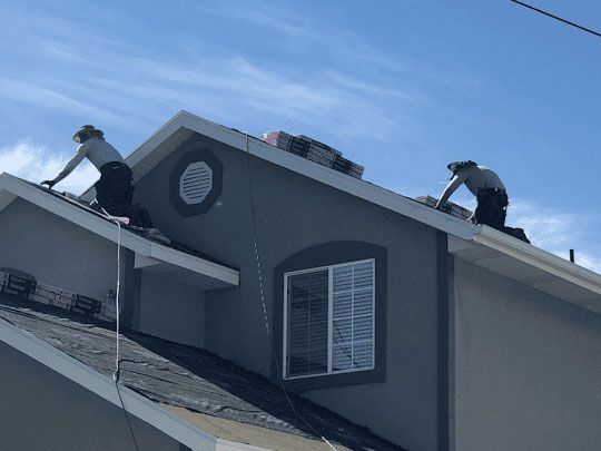 Two roofers on a gray house roof under a blue sky, working near the ridge.