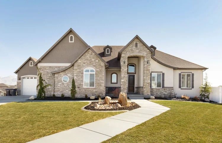 Suburban stone-and-stucco house with a front lawn, walkway, and landscaped entry under a clear sky