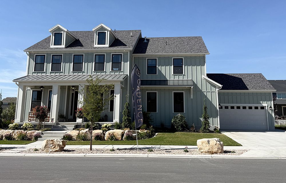 Large gray two-story house with a front porch, dormer windows, and a three-car garage on a sunny street.