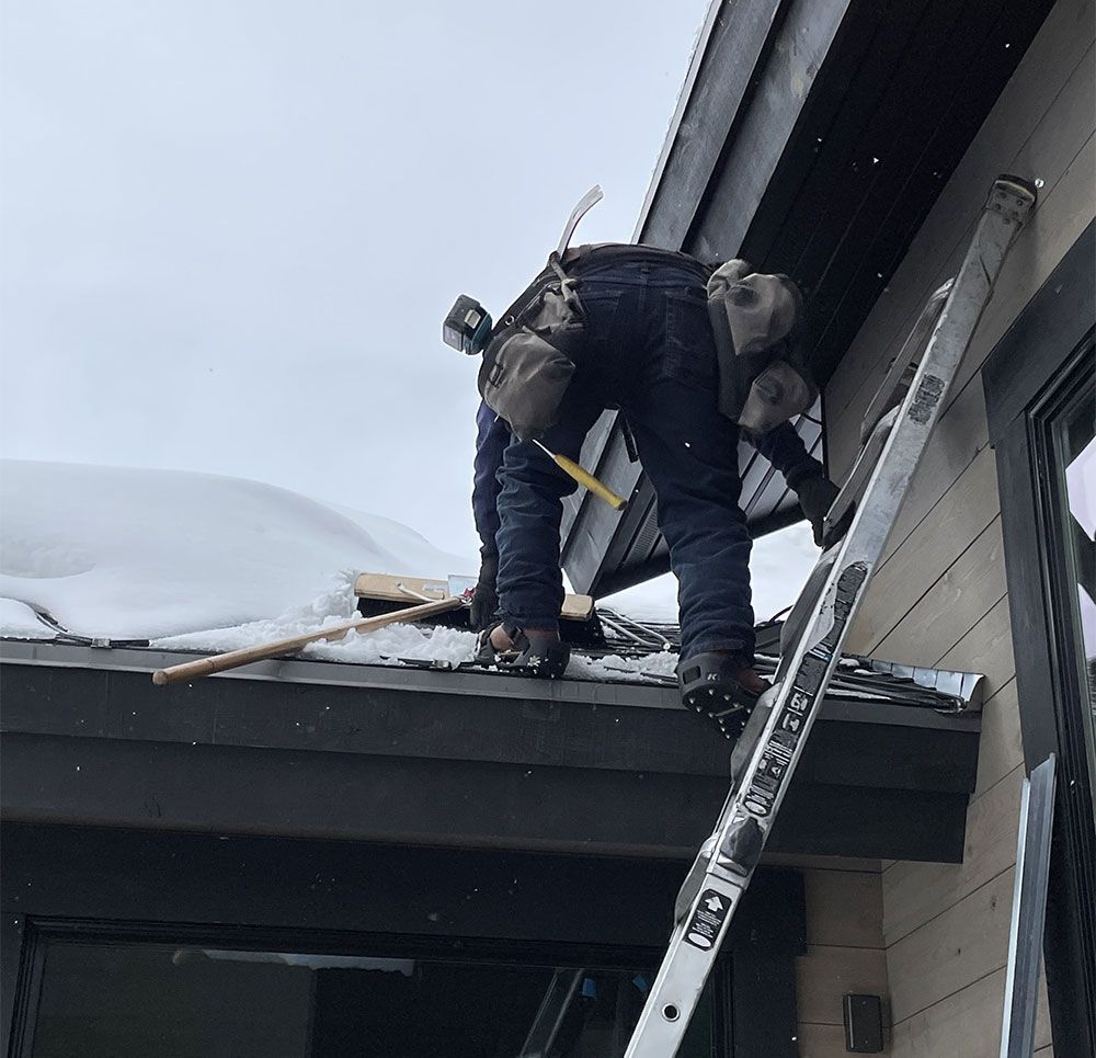 Worker clearing snow from a roof beside a ladder and house eaves