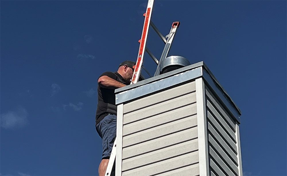 Worker on a rooftop using a ladder beside a white chimney under a clear blue sky