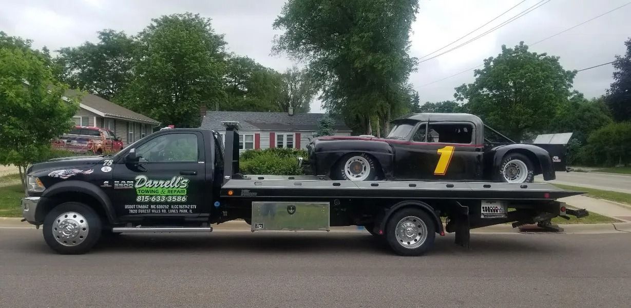 Tow truck hauling a black race car with number 1 on its side. Scene is outside, daytime.