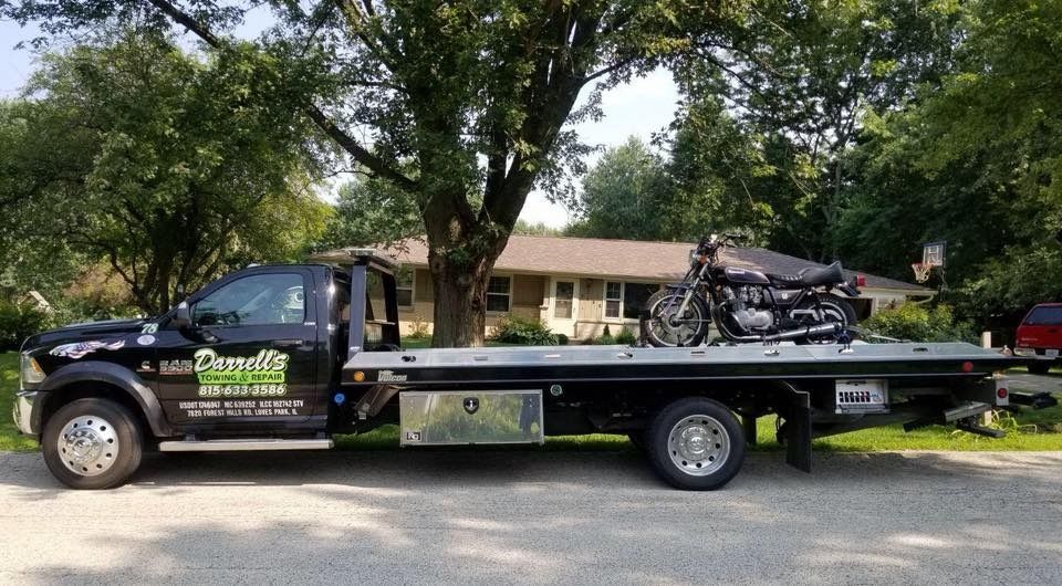Tow truck with a motorcycle on its flatbed in front of a house on a sunny day.