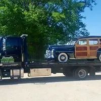 A black tow truck carrying a vintage wooden-paneled car on a sunny day.