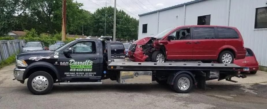 A red minivan severely damaged, being towed on a flatbed tow truck. The truck is black with green lettering.