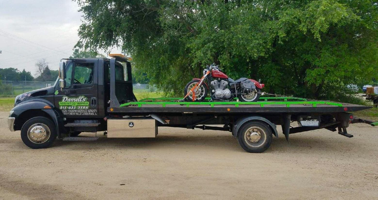 A black tow truck with a motorcycle on its flatbed, parked on gravel with trees in the background.