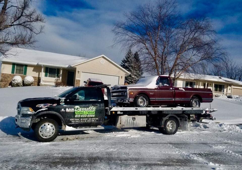 Tow truck carrying a maroon pickup truck on a snowy street in front of houses.