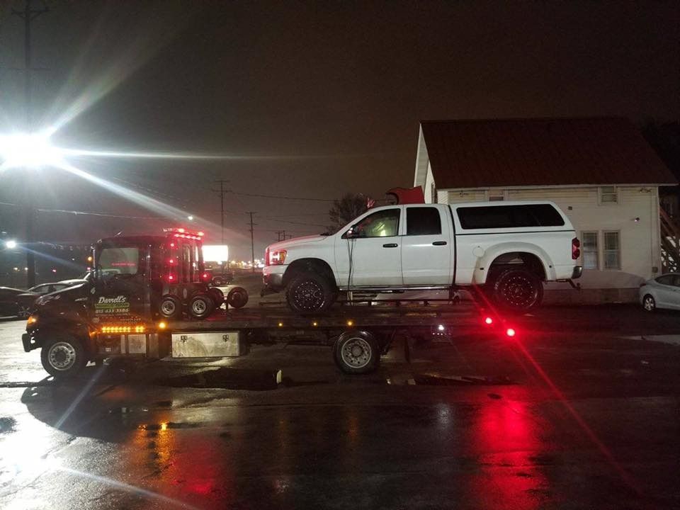 White pickup truck on a flatbed tow truck at night, lights reflecting on wet ground.