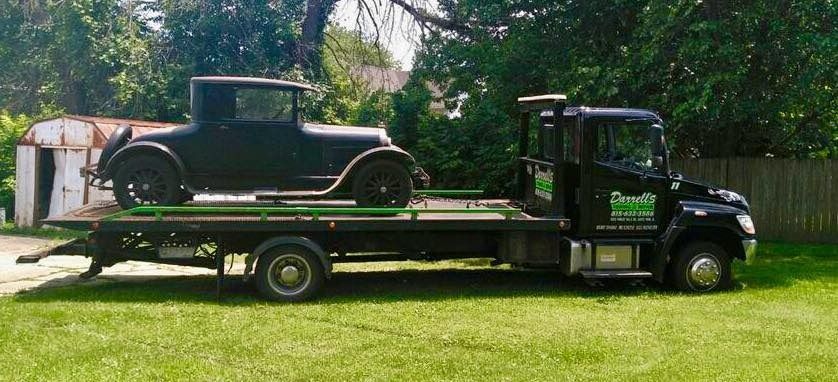 Black tow truck carrying a vintage black car on a green grassy lawn.